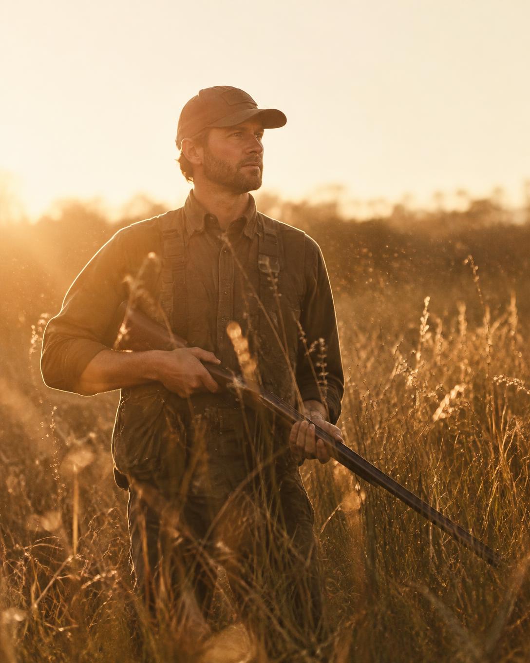Hunter carrying a shotgun through tall grass at golden hour